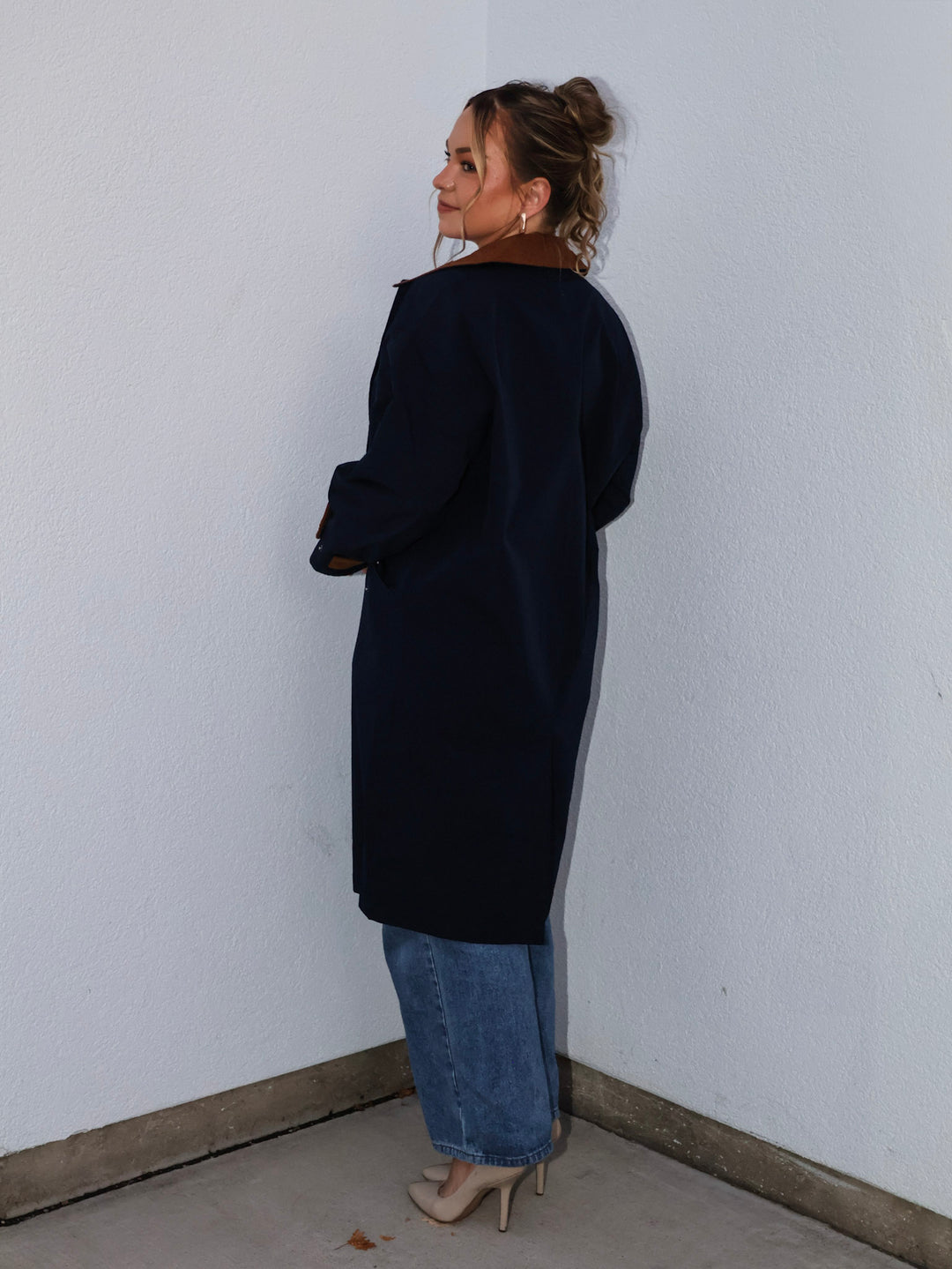 Woman wearing a black coat and blue jeans standing against a white wall.