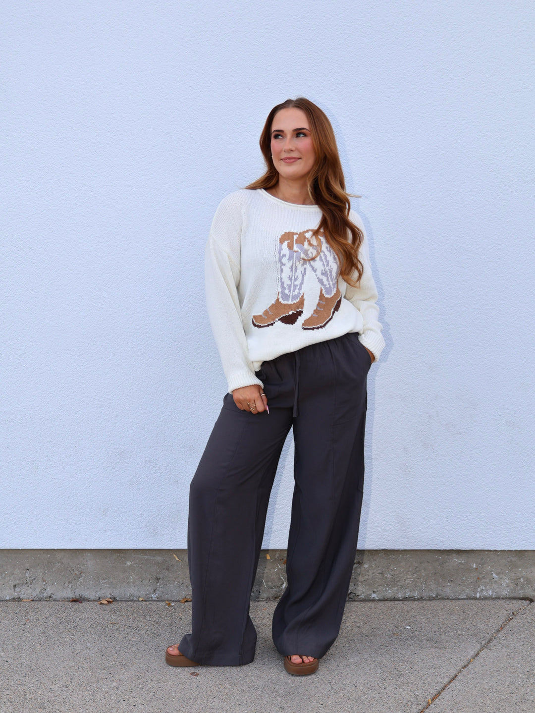 Woman wearing a white sweater with a graphic design and dark pants against a light blue wall.