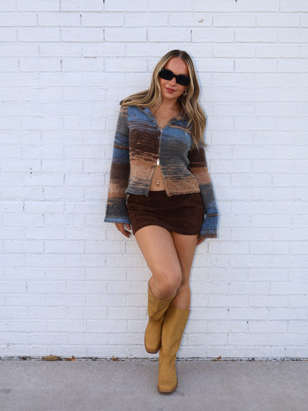 Woman wearing a patterned cardigan, brown shorts, and tan boots against a white brick wall.
