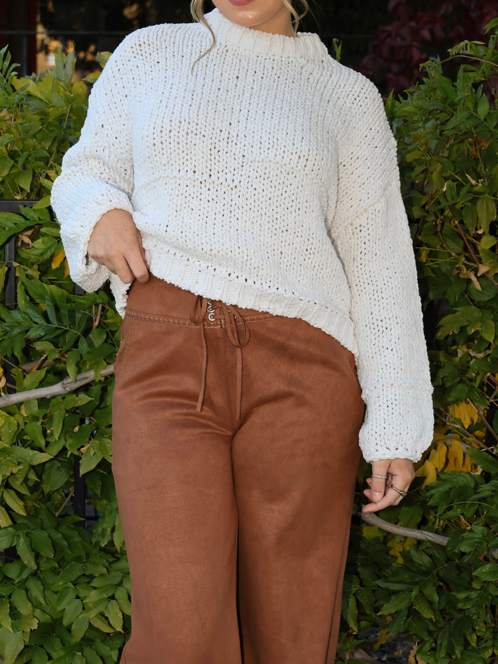 Person wearing a white knitted sweater and brown pants standing in front of green foliage.