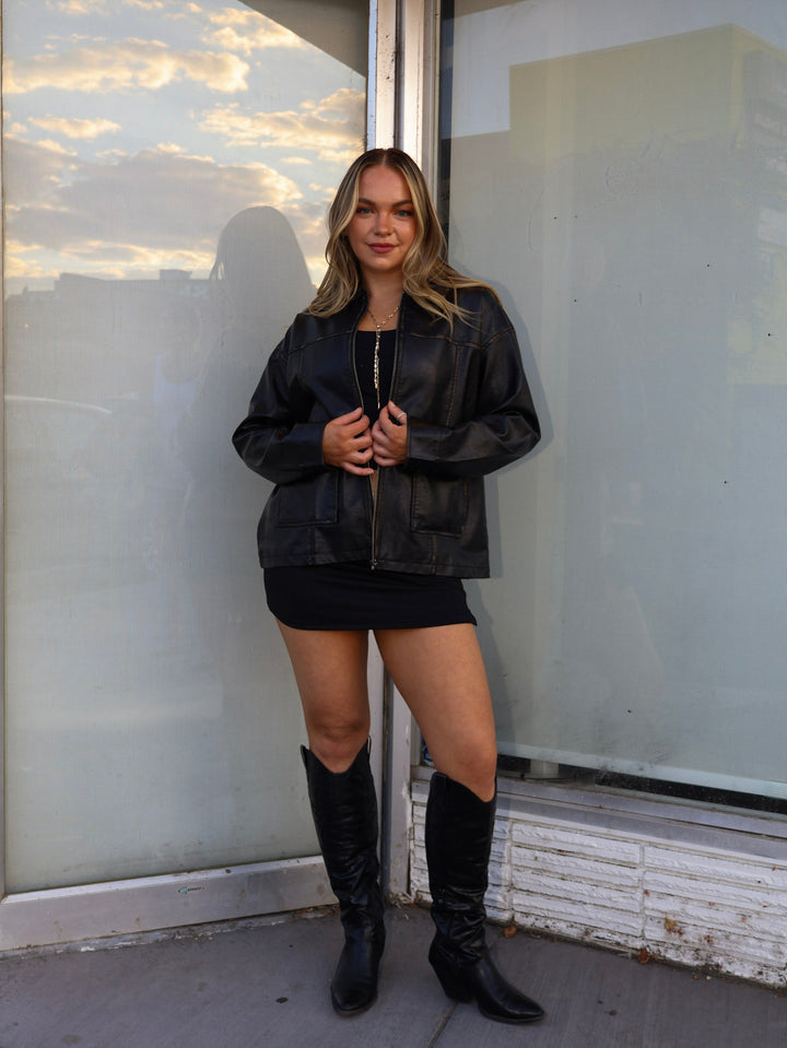 Woman wearing a black leather jacket, black skirt, and knee-high boots standing in front of a glass door.