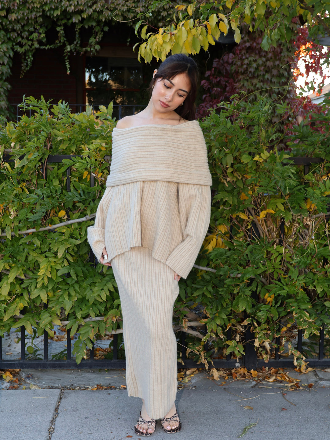 Woman wearing a beige off-shoulder top and wide-leg pants standing in front of green bushes.