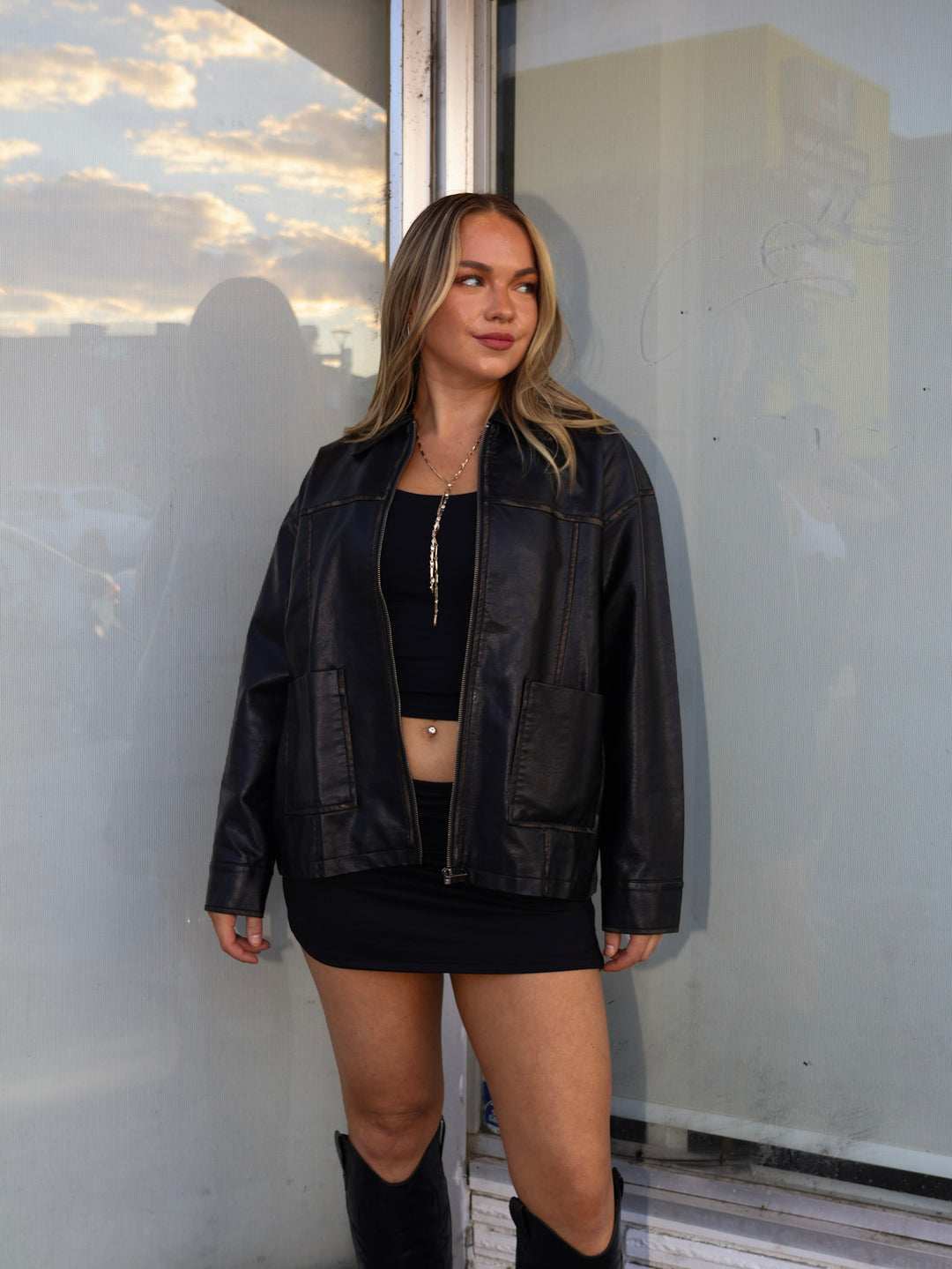 Woman wearing a black leather jacket and skirt standing against a reflective surface.