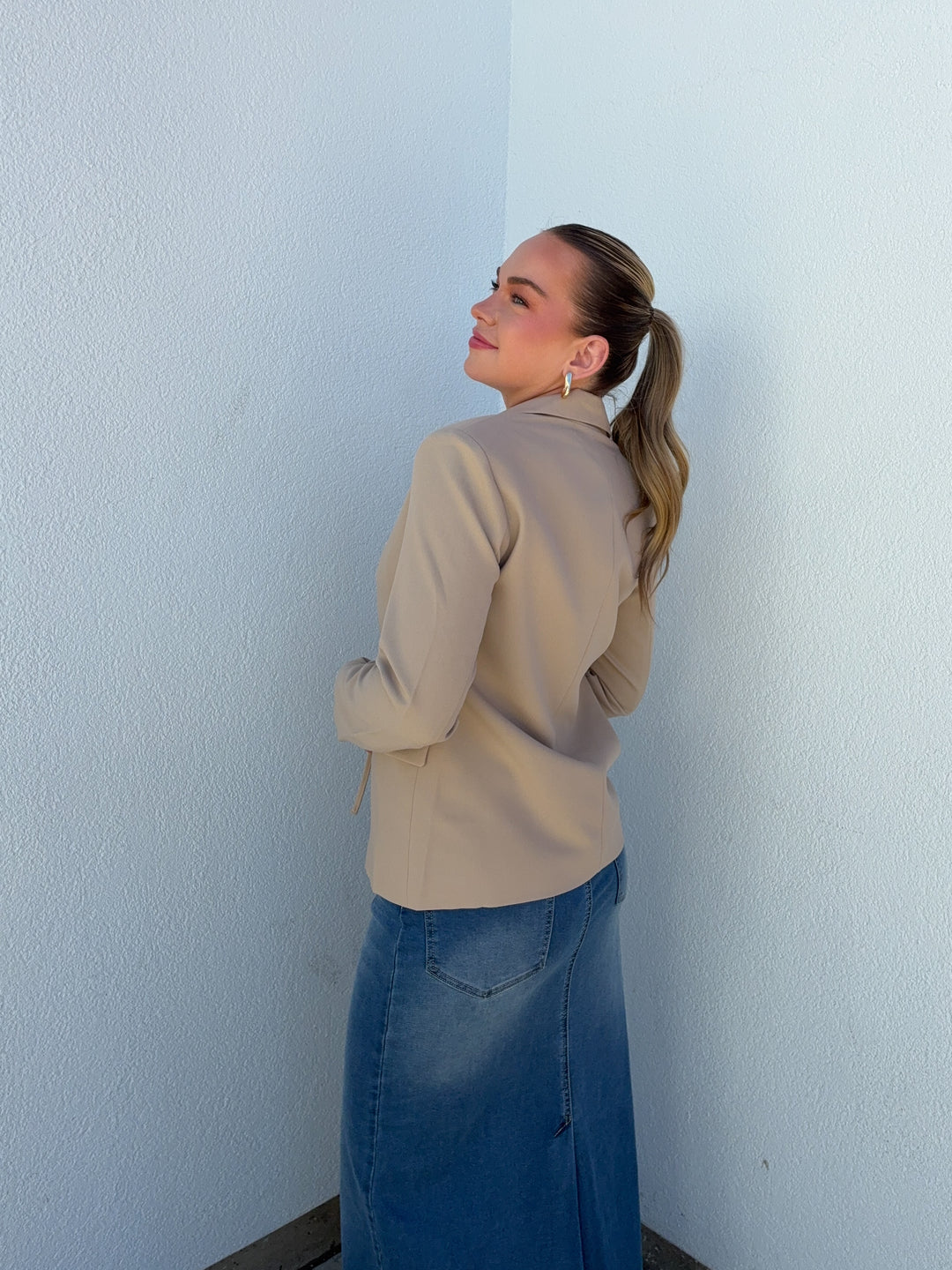Woman in beige jacket and blue skirt standing against a light blue wall