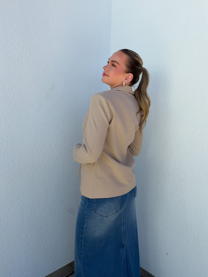 Woman in beige jacket and blue skirt standing against a light blue wall