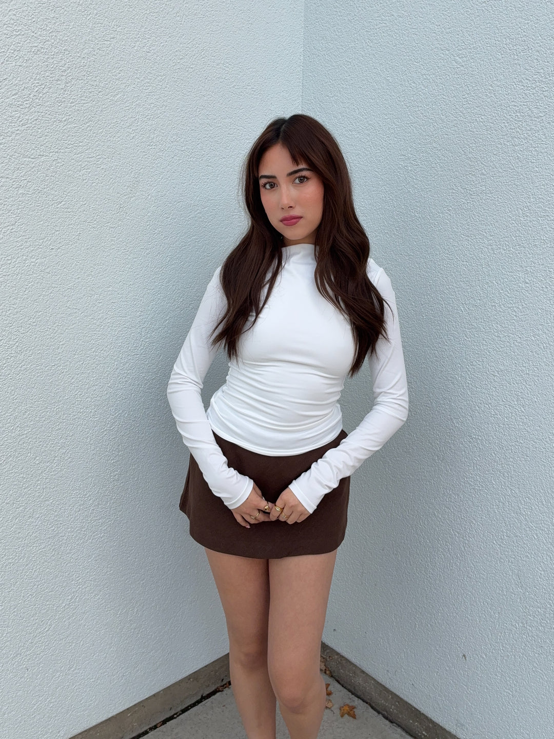 Woman wearing a white long-sleeve top and brown skirt against a light gray wall.