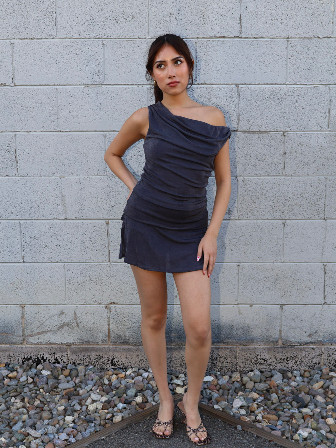 Woman wearing a one-shoulder dress standing against a gray brick wall.