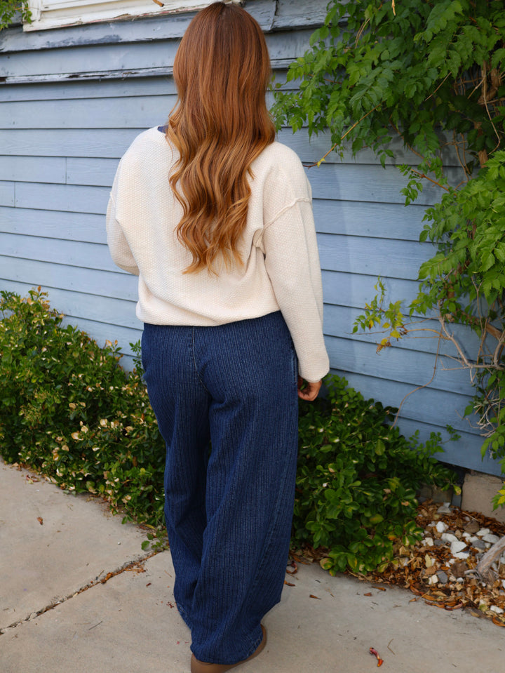 Person wearing a white sweater and blue pants standing in front of a blue house with greenery.