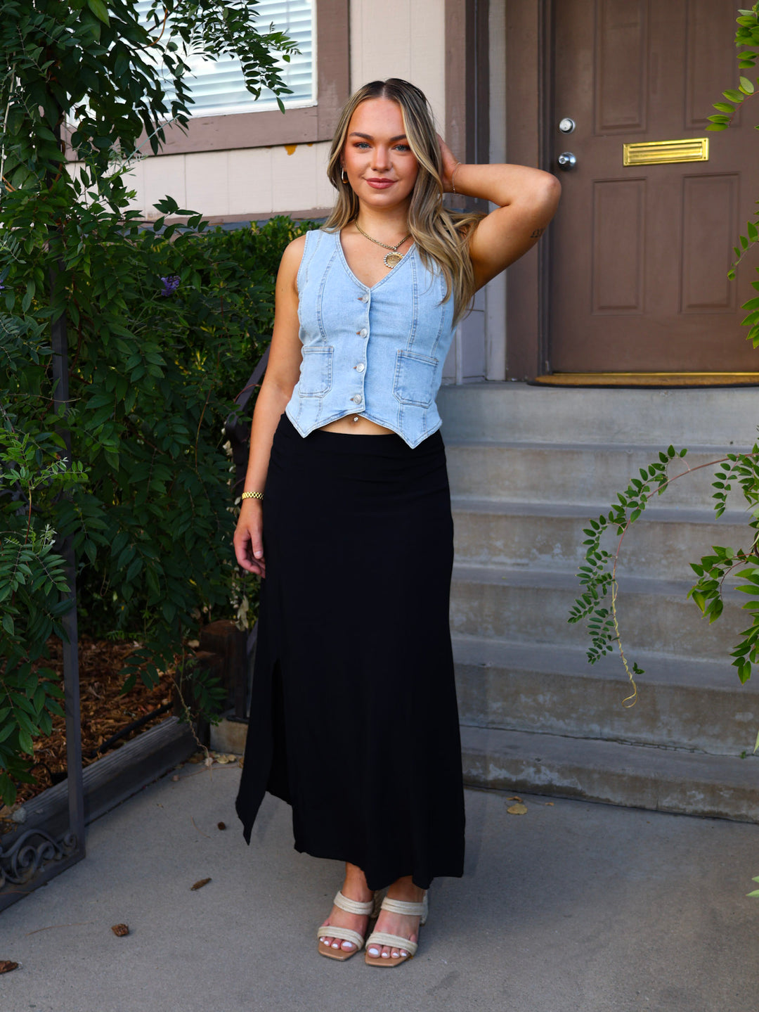 Woman standing on a porch wearing a light blue denim vest over a black top and black skirt.