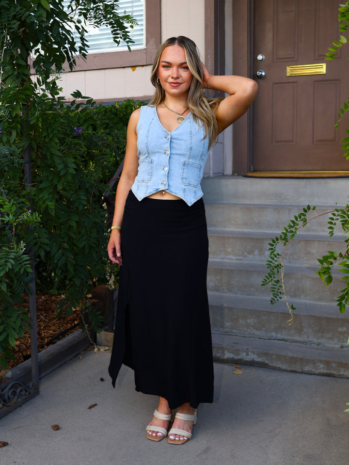 Woman standing on a porch wearing a light blue denim vest over a black top and black skirt.