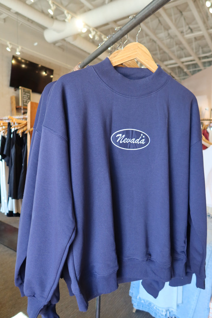 A navy blue crewneck sweatshirt hanging on a wooden hanger on a clothing rack inside a retail store. The sweatshirt features a white oval 'Nevada' logo printed on the center chest.