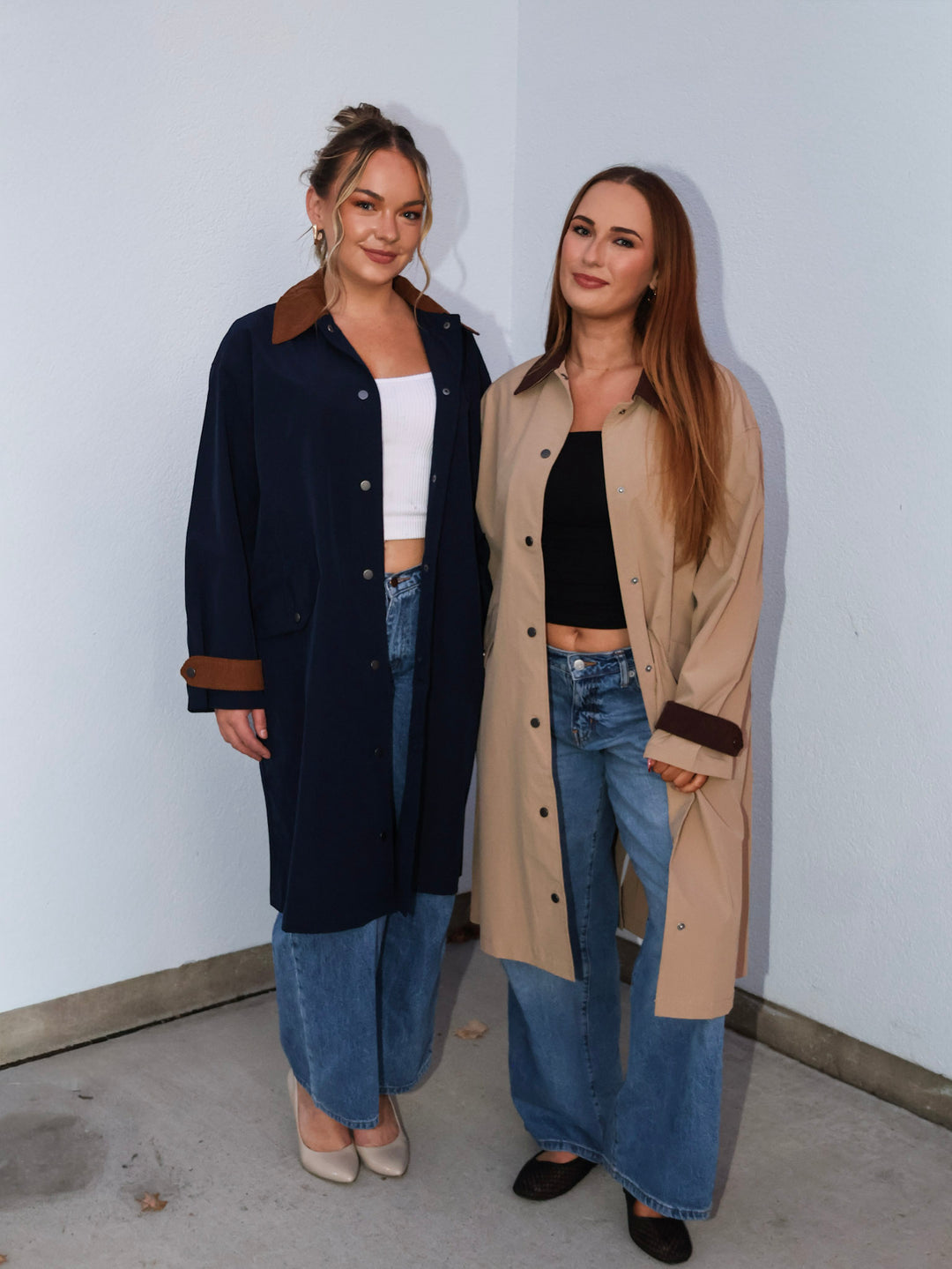 Two women standing side by side wearing long coats and jeans against a plain wall.