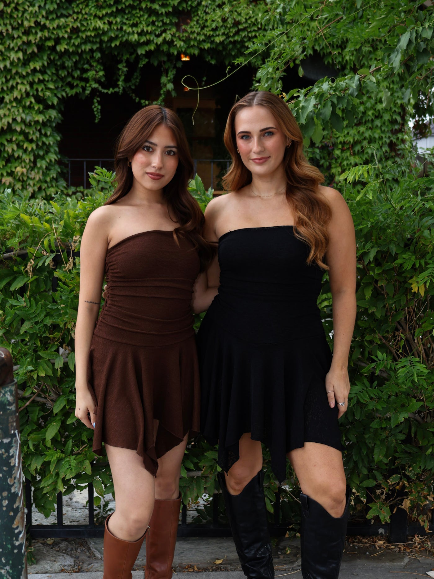 Two women wearing strapless dresses standing in front of a green hedge.