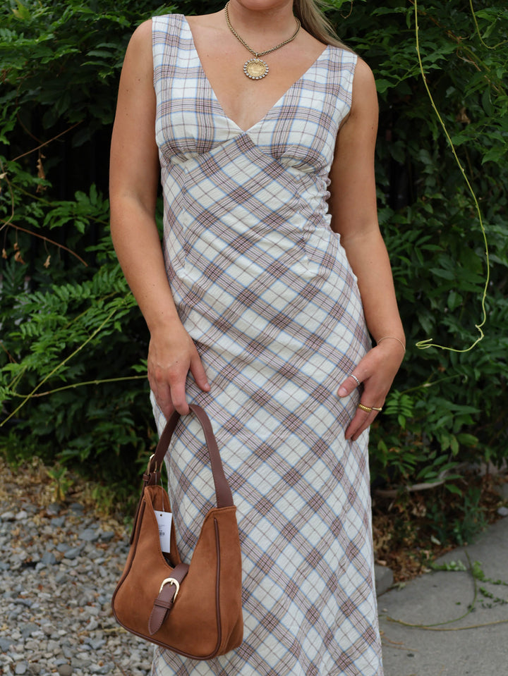 Person wearing a plaid dress holding a brown handbag against a green foliage background