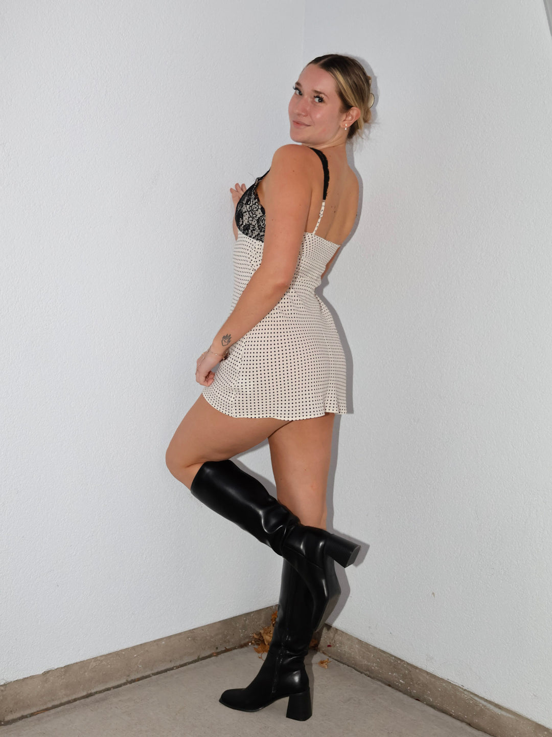 Woman wearing a white dress with black lace details and black knee-high boots against a plain wall.