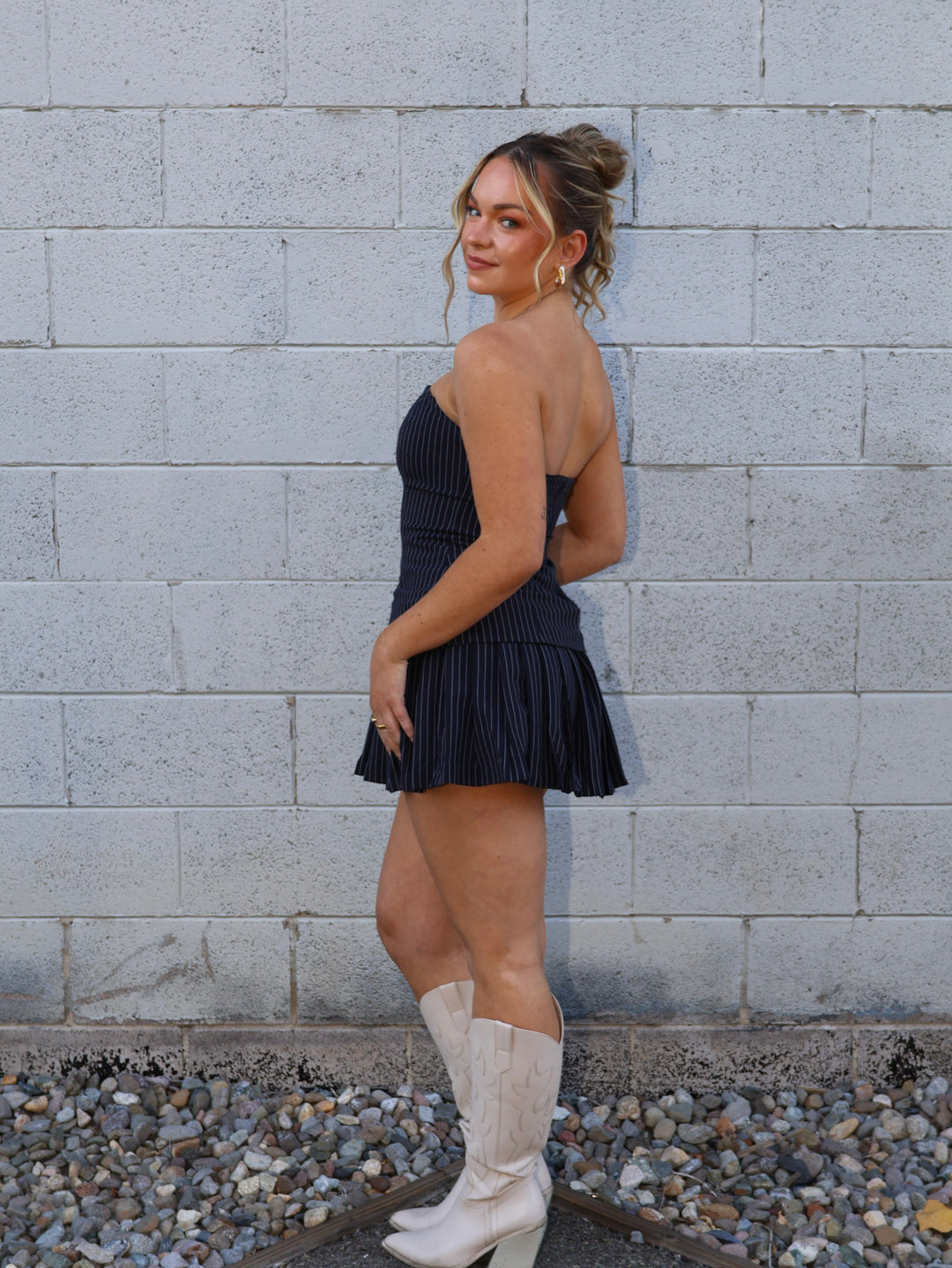 Woman in a navy dress and white boots standing against a gray brick wall.