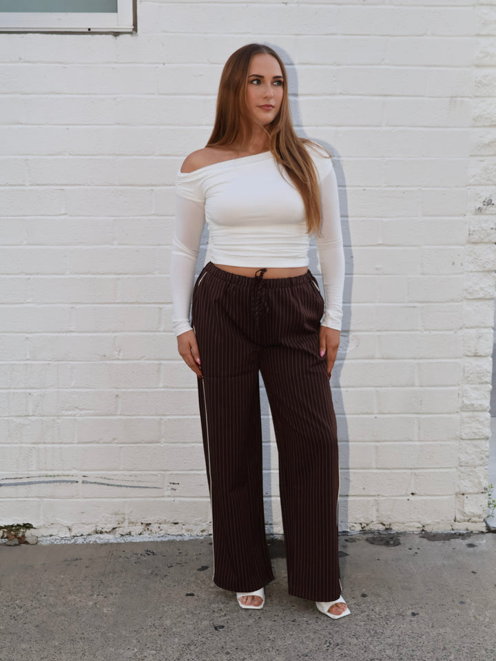 Woman wearing a white off-shoulder top and brown pinstripe pants against a white brick wall.