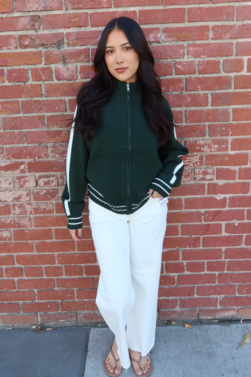 A woman stands against a red brick wall wearing a dark green zip-up track jacket with white sleeve stripes, white wide-leg pants, and brown sandals.
