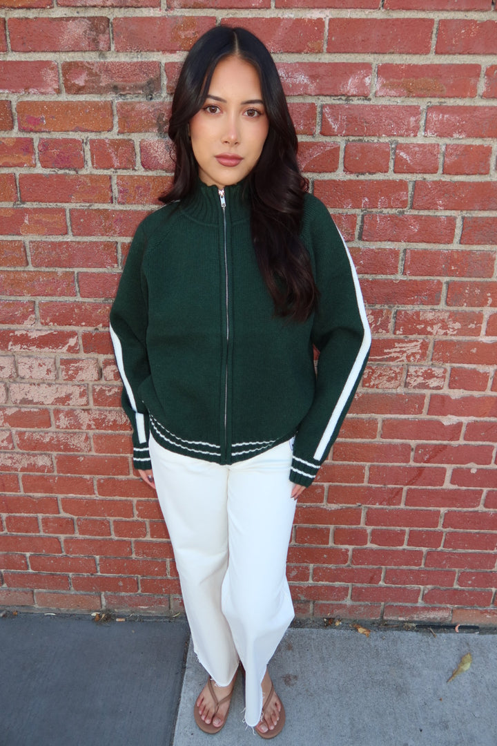 A woman stands in front of a red brick wall wearing a dark green zip-up track jacket with white stripes on the sleeves and white pants.