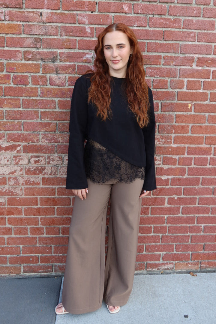 Model in the black Charlie asymmetric lace trim top and taupe Billie tailored wide leg trousers by a red brick wall