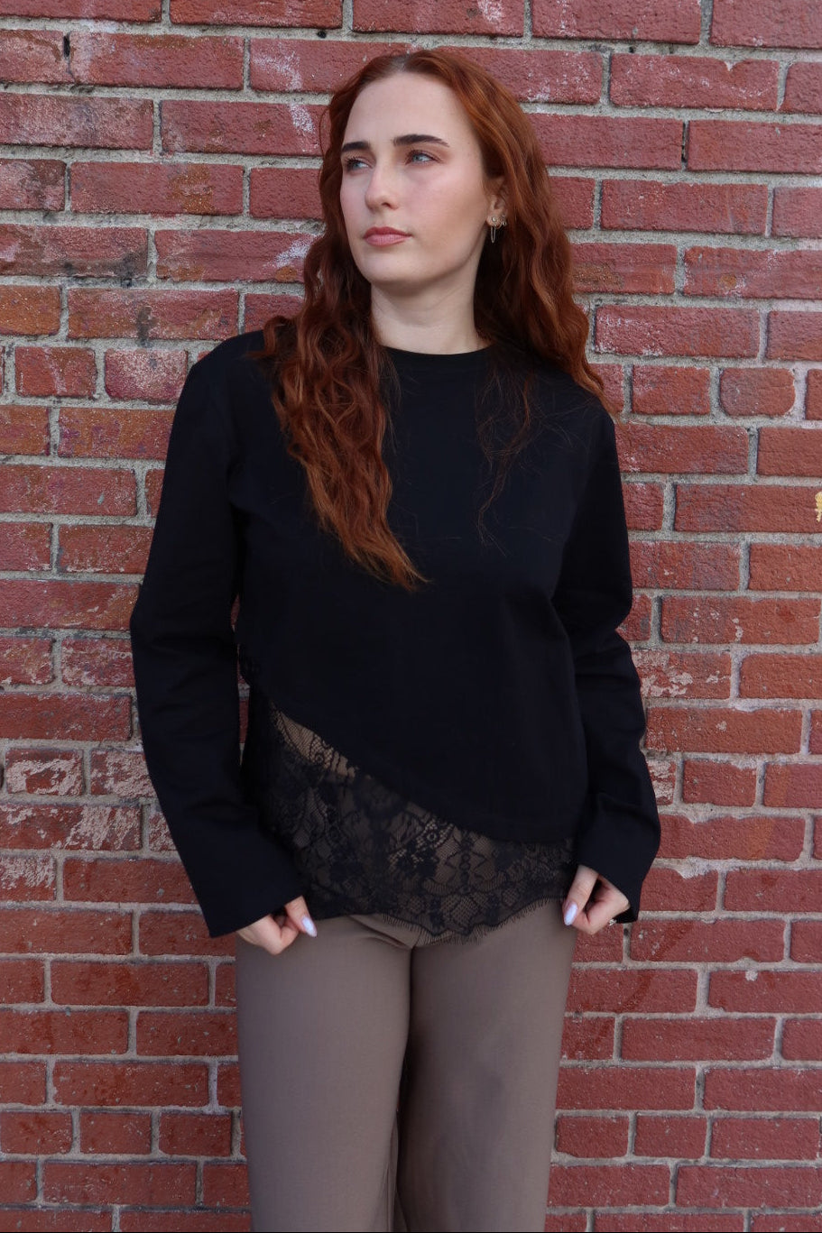 Close view of the long sleeve black Charlie asymmetric lace trim top, hands in pockets, against a brick wall
