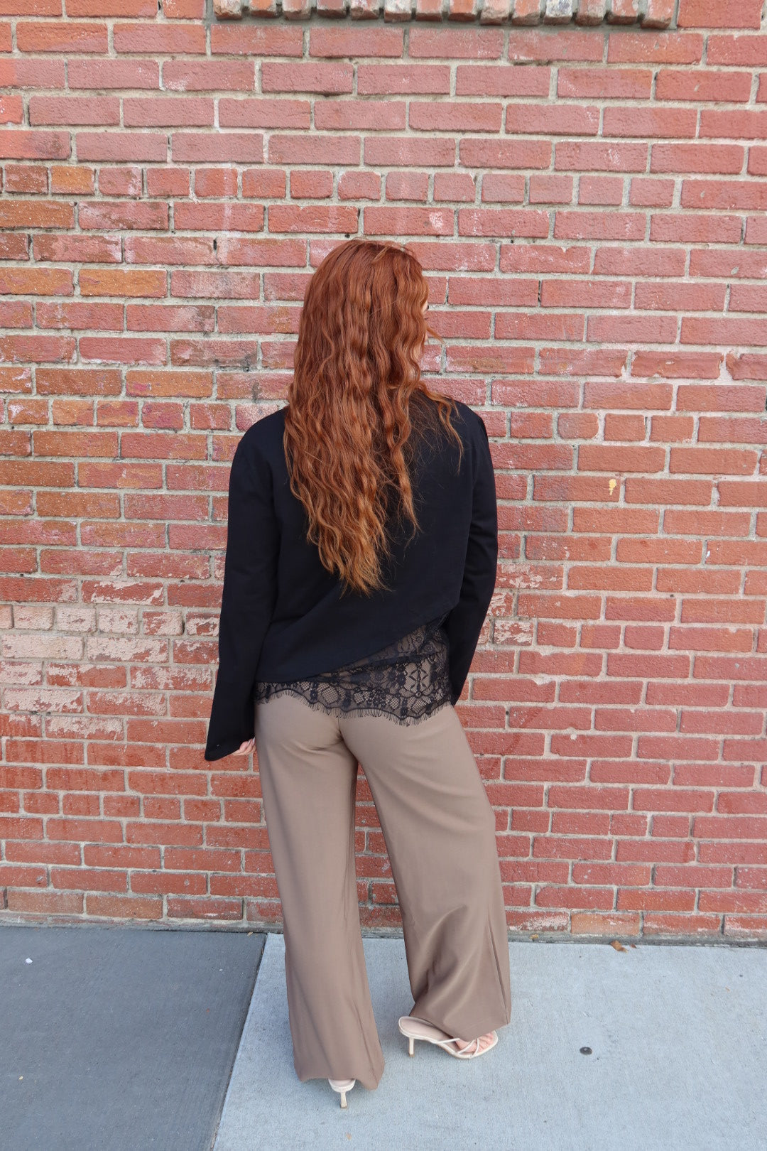 Back view of the taupe Billie tailored wide leg trousers with a black lace trim top, against a brick wall