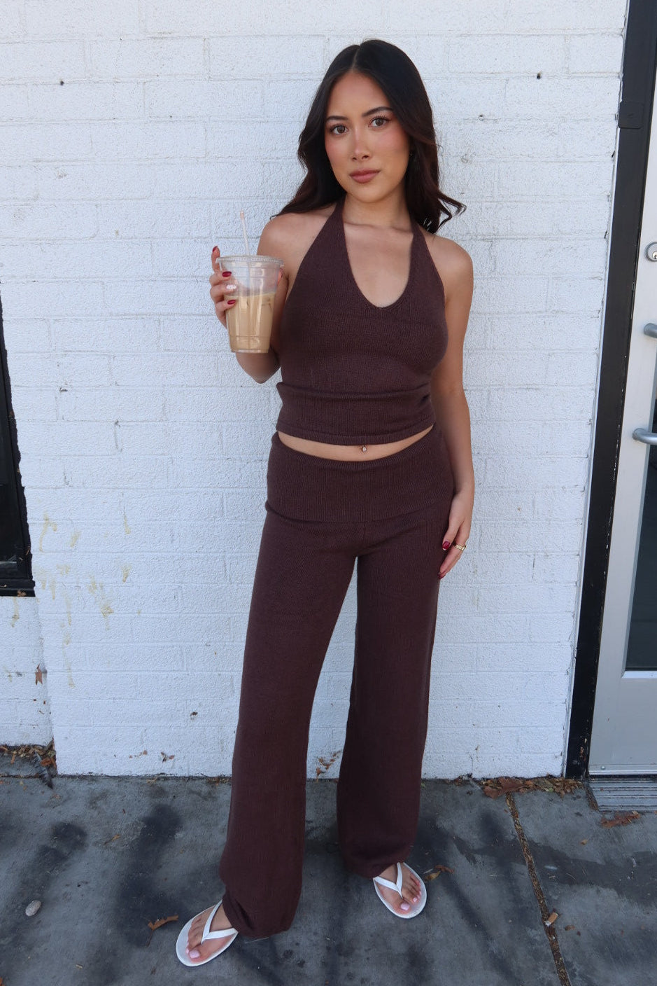 Model in the brown Kelsey knit halter top and trouser set with white flip flops, holding an iced coffee by a white brick wall