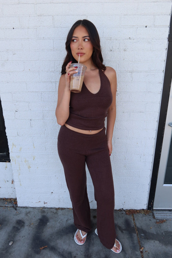 Model sipping an iced coffee in the brown Kelsey knit halter top and trouser set, front view by a white brick wall