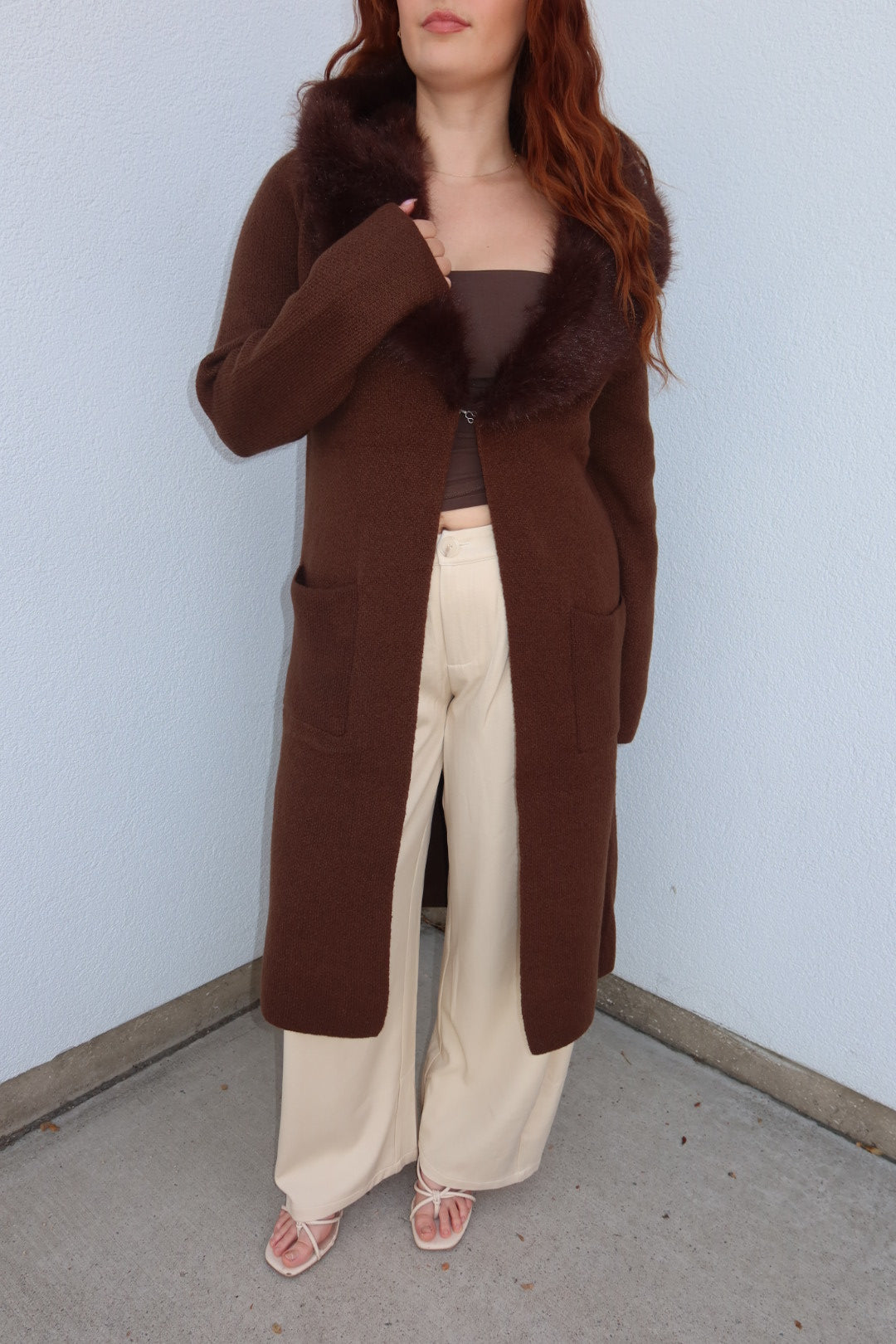 Model wearing the long brown Felicity faux fur collar cardigan over a brown top and cream wide leg pants, front view
