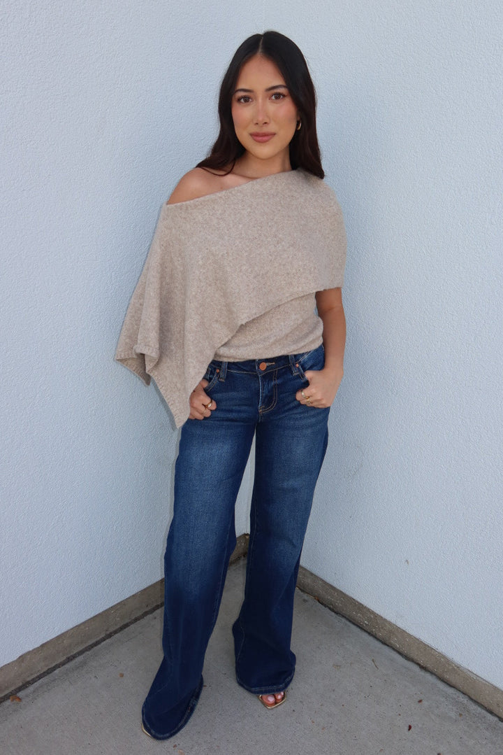 Model wearing the oatmeal Ainsley off shoulder asymmetric knit poncho sweater with dark wash flare jeans, full length