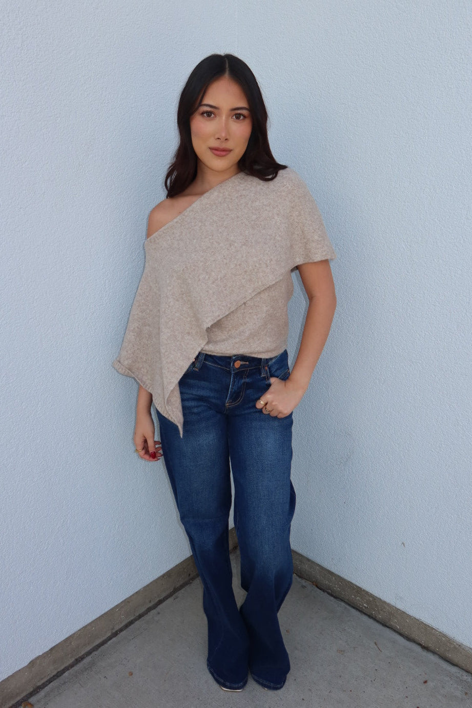 Model in the oatmeal Ainsley off shoulder asymmetric knit poncho sweater and dark wash jeans, hands in pockets
