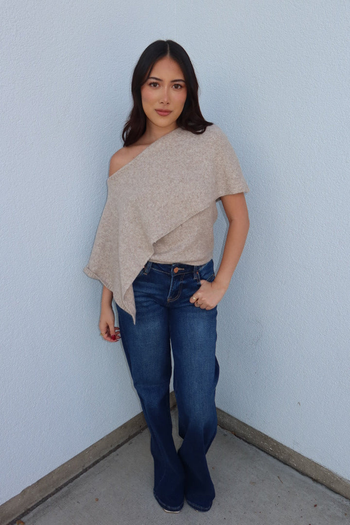 Model in the oatmeal Ainsley off shoulder asymmetric knit poncho sweater and dark wash jeans, hands in pockets