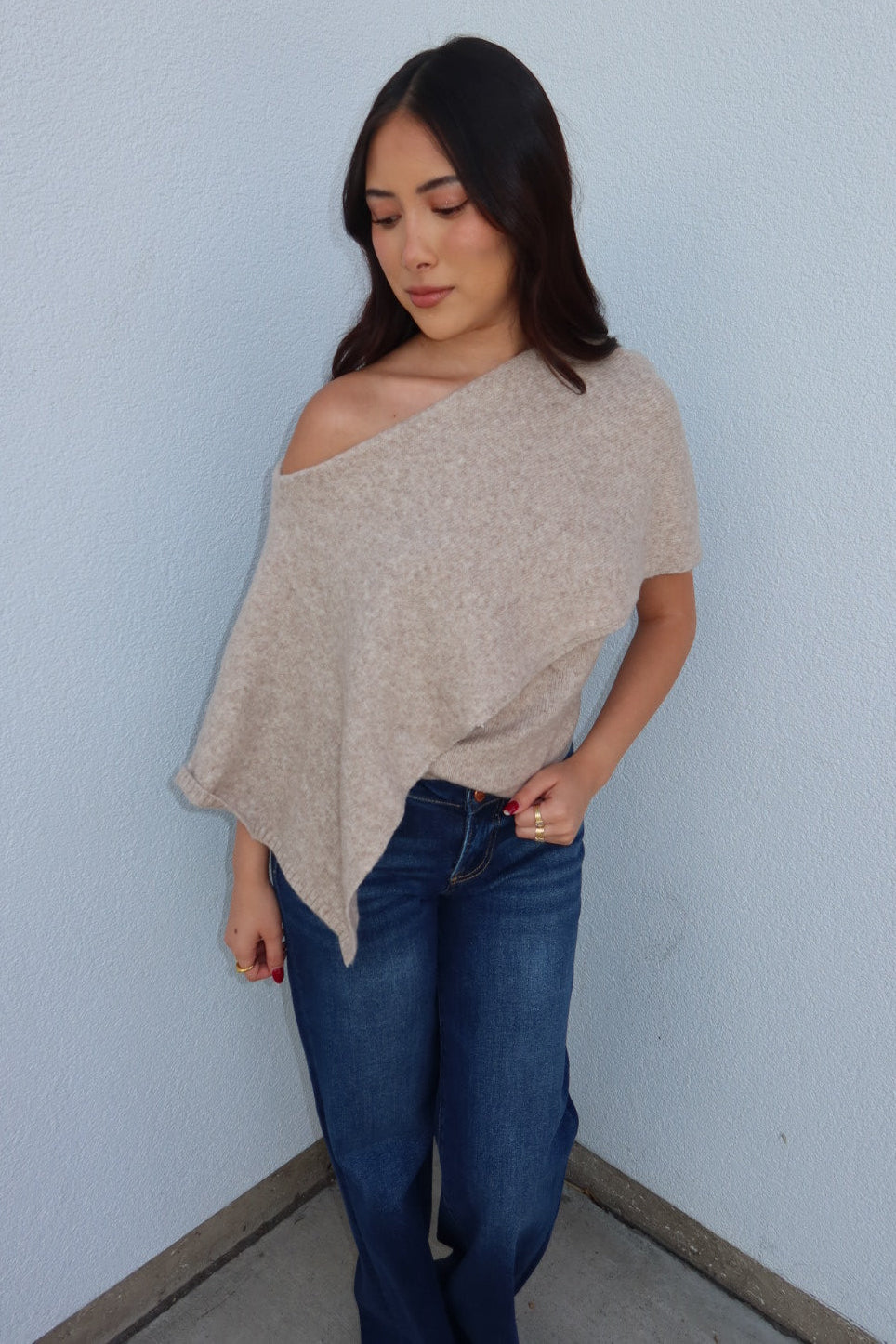 Model wearing the oatmeal Ainsley off shoulder asymmetric knit poncho sweater with dark jeans, looking down, front view