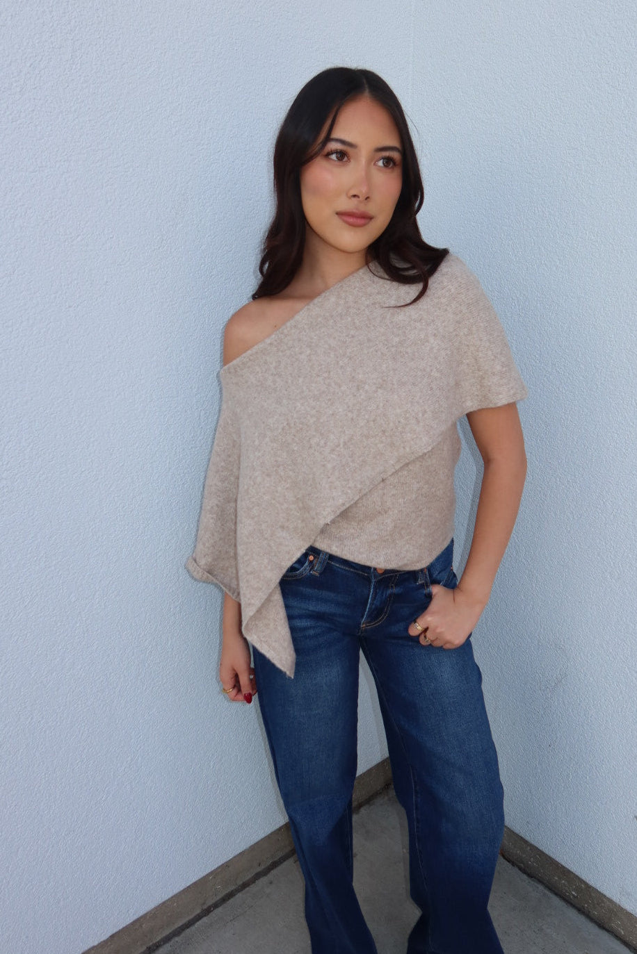 Model in the oatmeal Ainsley off shoulder asymmetric knit poncho sweater, hand in pocket, worn with dark wash jeans