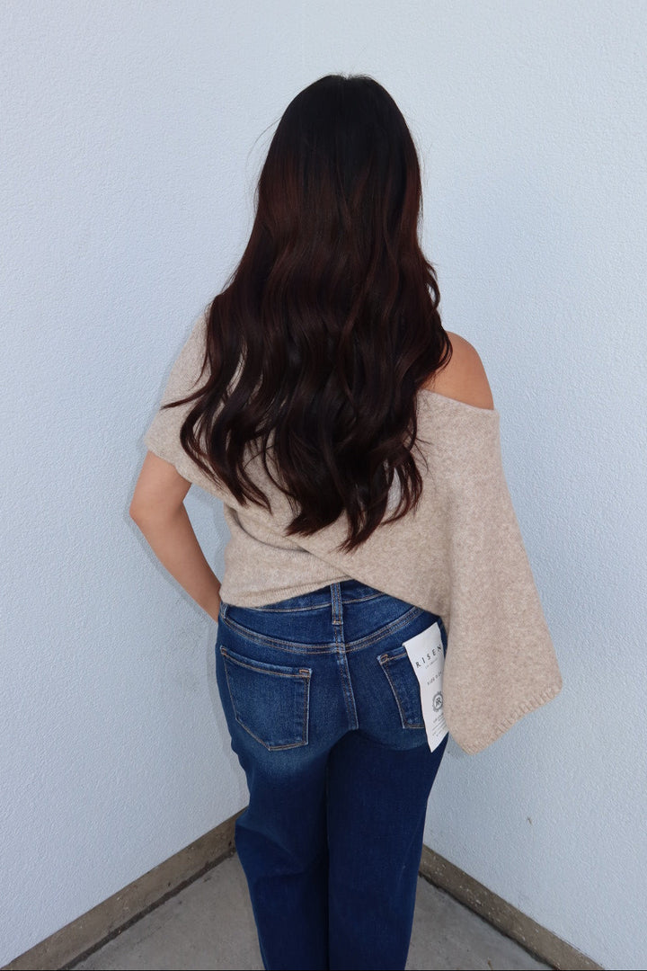 Back view of the oatmeal Ainsley off shoulder asymmetric knit poncho sweater with dark wash jeans