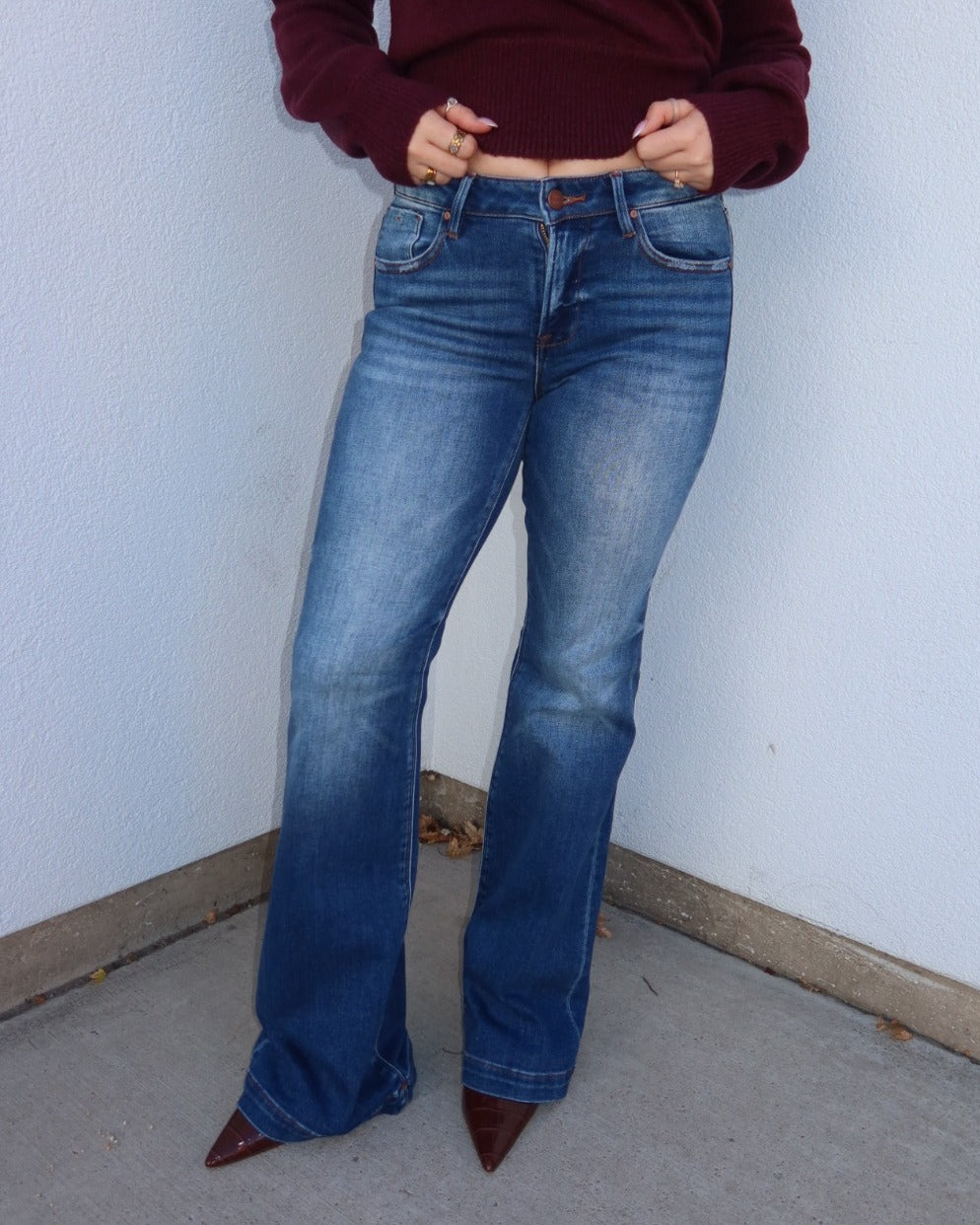 A person wearing medium-wash blue flare jeans with a maroon cropped long-sleeve top and brown pointed-toe heels, standing against a light gray wall.