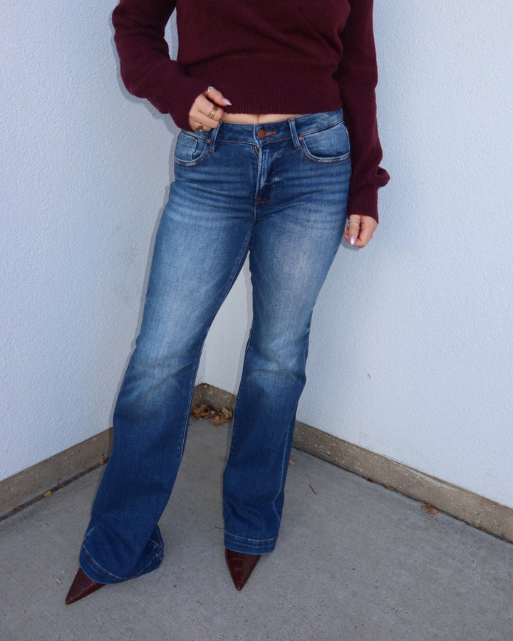 A person wearing medium-wash blue flare jeans with a maroon long-sleeve top and brown pointed-toe heels, standing against a light gray wall.
