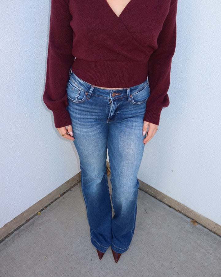 A person wearing a burgundy wrap-style knit sweater and medium-wash blue jeans, standing against a light blue wall in a corner.
