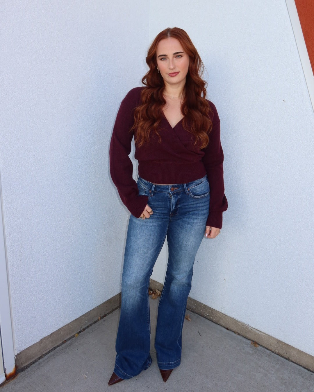 A woman with long wavy red hair stands in a corner wearing a dark burgundy wrap-style long-sleeve top, blue flare jeans, and brown pointed-toe heels against a white wall.