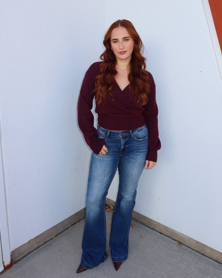 A woman with long wavy red hair stands in a corner wearing a dark burgundy wrap-style long-sleeve top, blue flare jeans, and brown pointed-toe heels against a white wall.